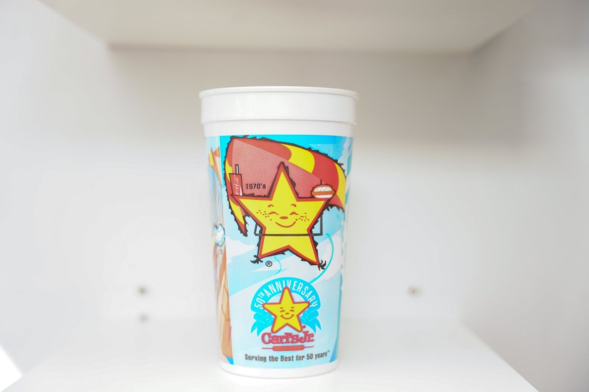 Carl's Jr. 50th Anniversary white plastic cup with Happy Star mascot, 1970s restaurant illustration, and Serving the Best for 50 years tagline