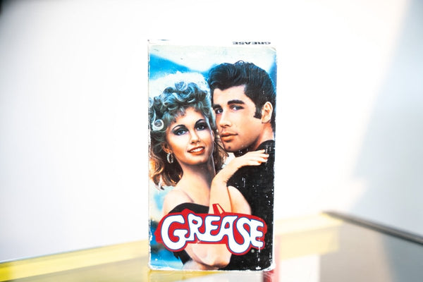 Grease VHS blue sleeve with Olivia Newton-John Sandy perm and John Travolta Danny Zuko leather jacket close-up red yellow chrome GREASE logo