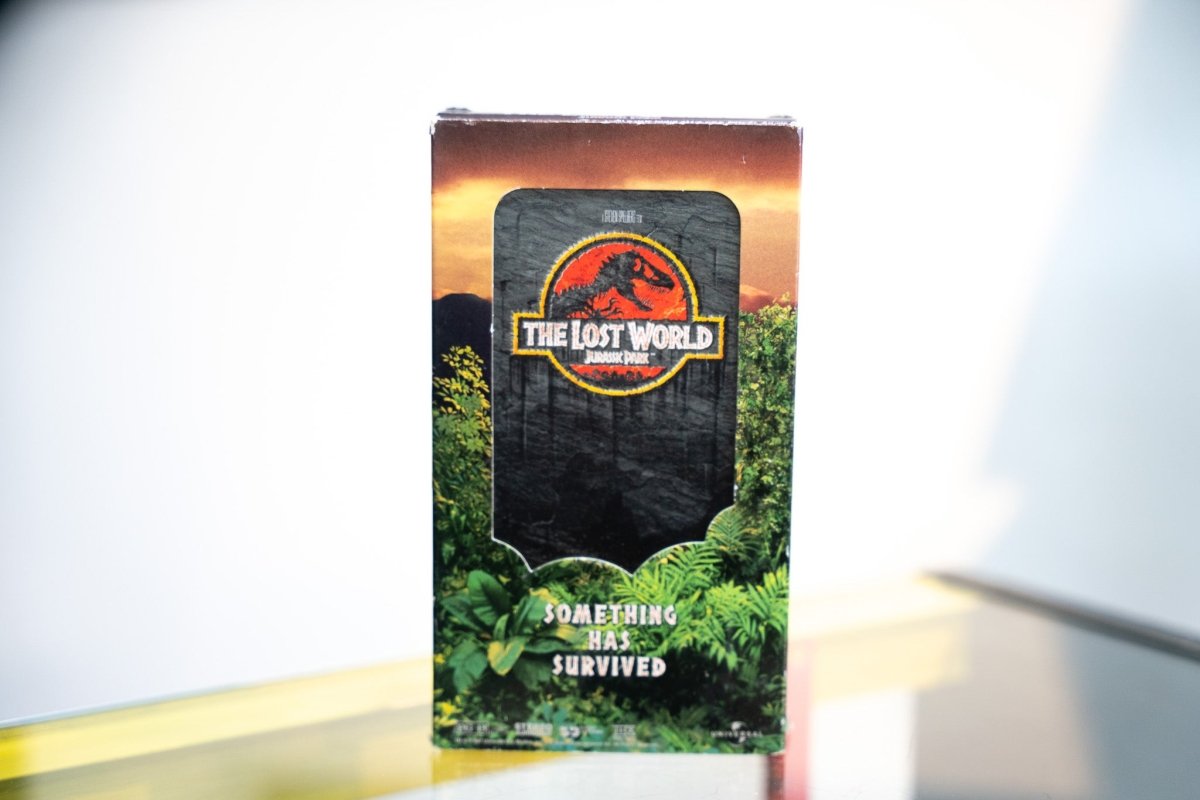 The Lost World Jurassic Park VHS sleeve with jungle ferns sunset volcanic mountains dark stone tablet red gold T-Rex logo Something Has Survived