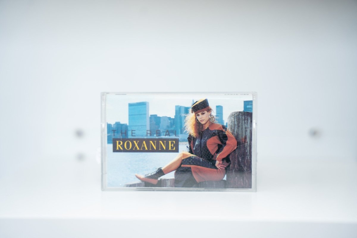 The Real Roxanne cassette tape clear case Roxanne on NYC waterfront dock pilings Manhattan skyline orange black outfit THE REAL ROXANNE text