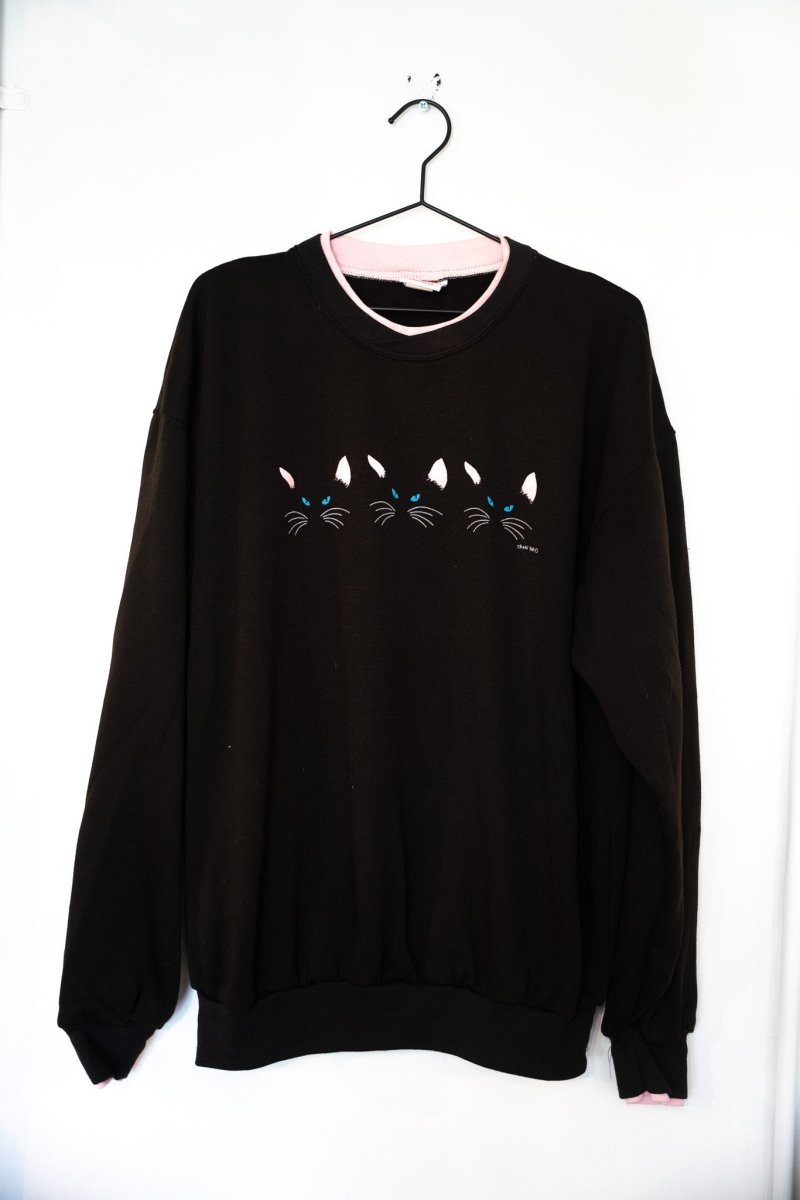 Black 1985 Kitty Boy Eyes and Ears Acton sweatshirt with three pairs of blue cat eyes and ears peeking up and pink collar trim