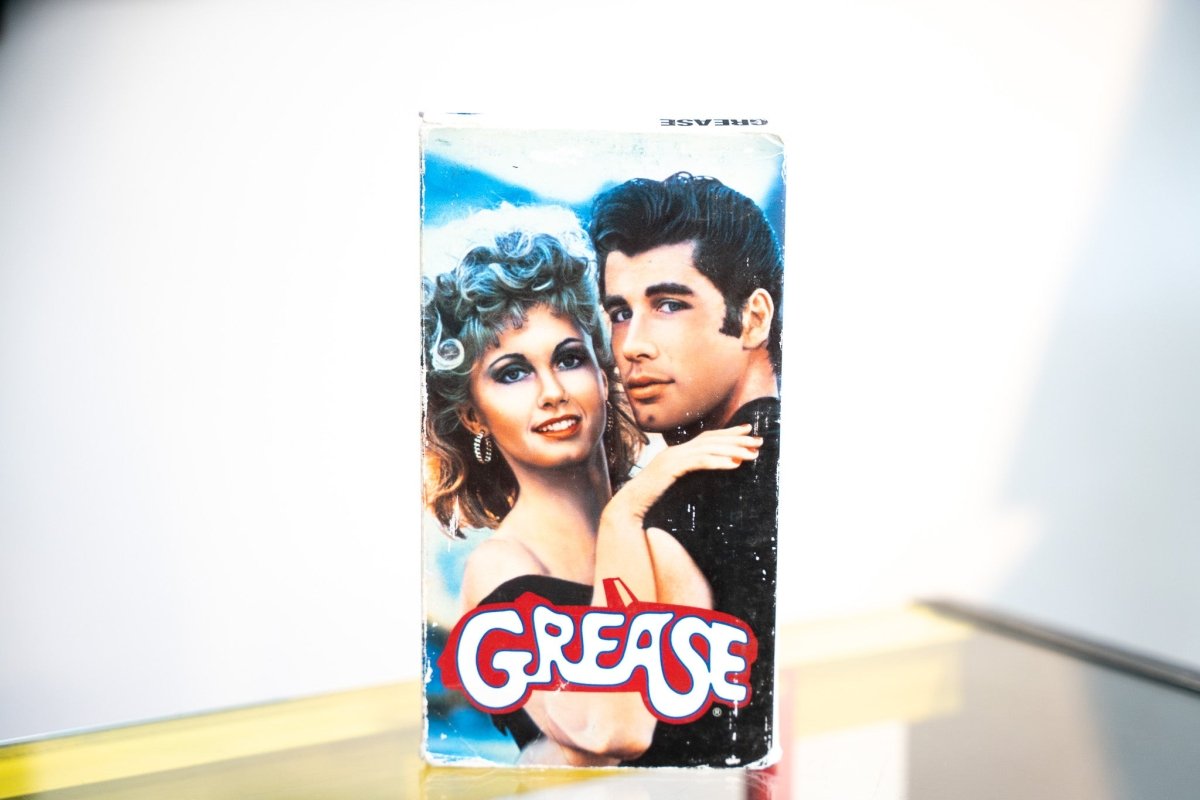 Grease VHS blue sleeve with Olivia Newton-John Sandy perm and John Travolta Danny Zuko leather jacket close-up red yellow chrome GREASE logo