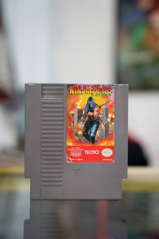 Nintendo NES Ninja Gaiden gray cartridge with Ryu Hayabusa ninja crouching on rooftop against fiery orange cityscape with yellow and red title